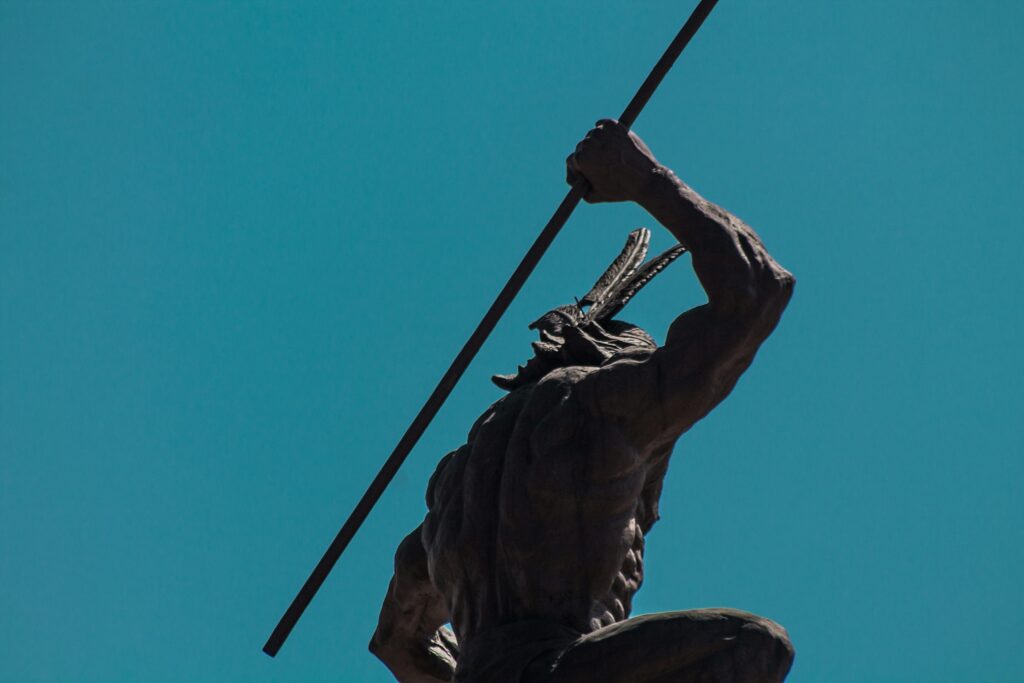 Low angle shot of an indigenous warrior statue with spear under a clear blue sky in a dynamic pose.