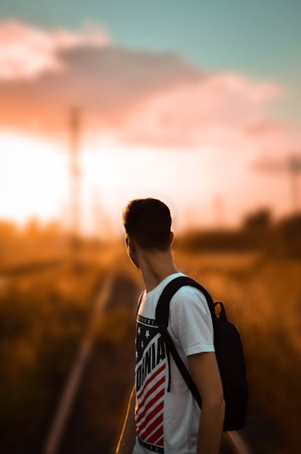 A young man stands on railway tracks at sunrise, wearing a backpack and white shirt.