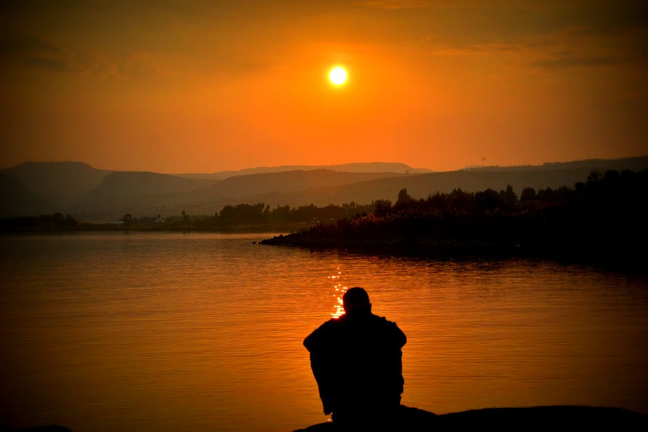 A person sits alone by a tranquil lake, with a stunning sunset in the background.