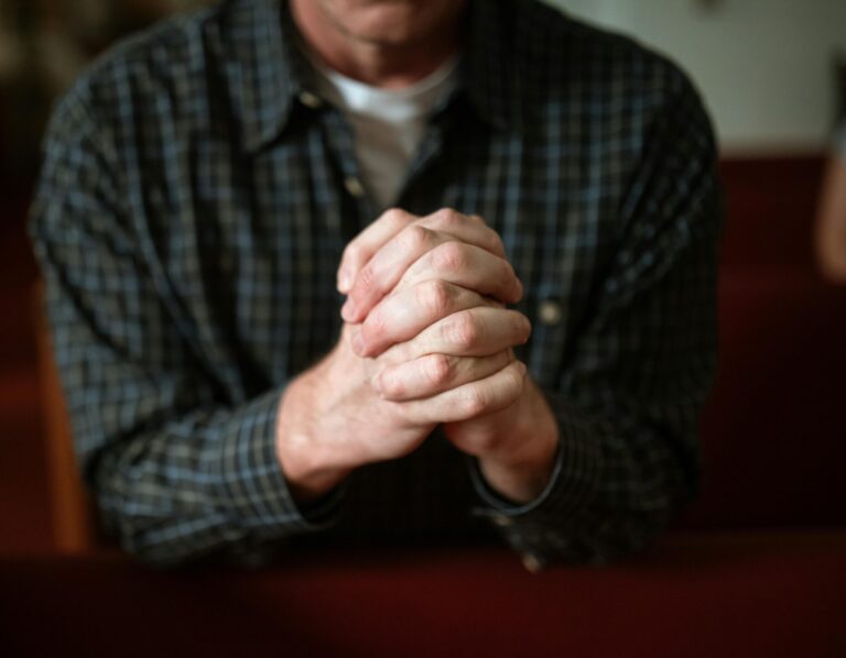 A man in a checked shirt praying in a church, hands clasped together.